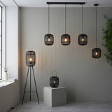 Bracken Pendant Light in black rattan and metal, shown as multiple cylindrical cage pendants and a matching table lamp in a grey-walled living space.