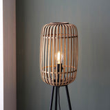 Bracken Floor Lamp in black and natural, showing a cylindrical bamboo cage shade with visible bulb on a black tripod base, set against a grey wall.