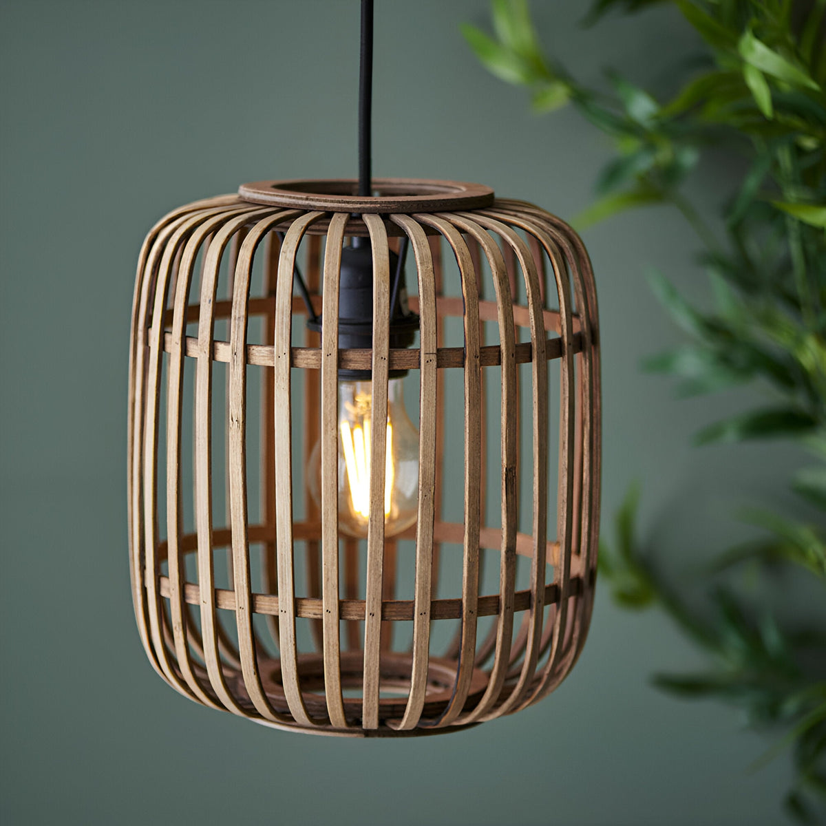 Endon Bracken pendant light with a natural bamboo cage shade and matt black flex, lit by an E27 filament bulb against a green backdrop with foliage.