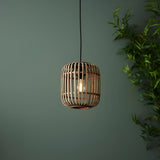 Endon Bracken pendant light with natural bamboo cage shade and matt black flex, lit E27 bulb, hung against a green wall beside bamboo foliage.