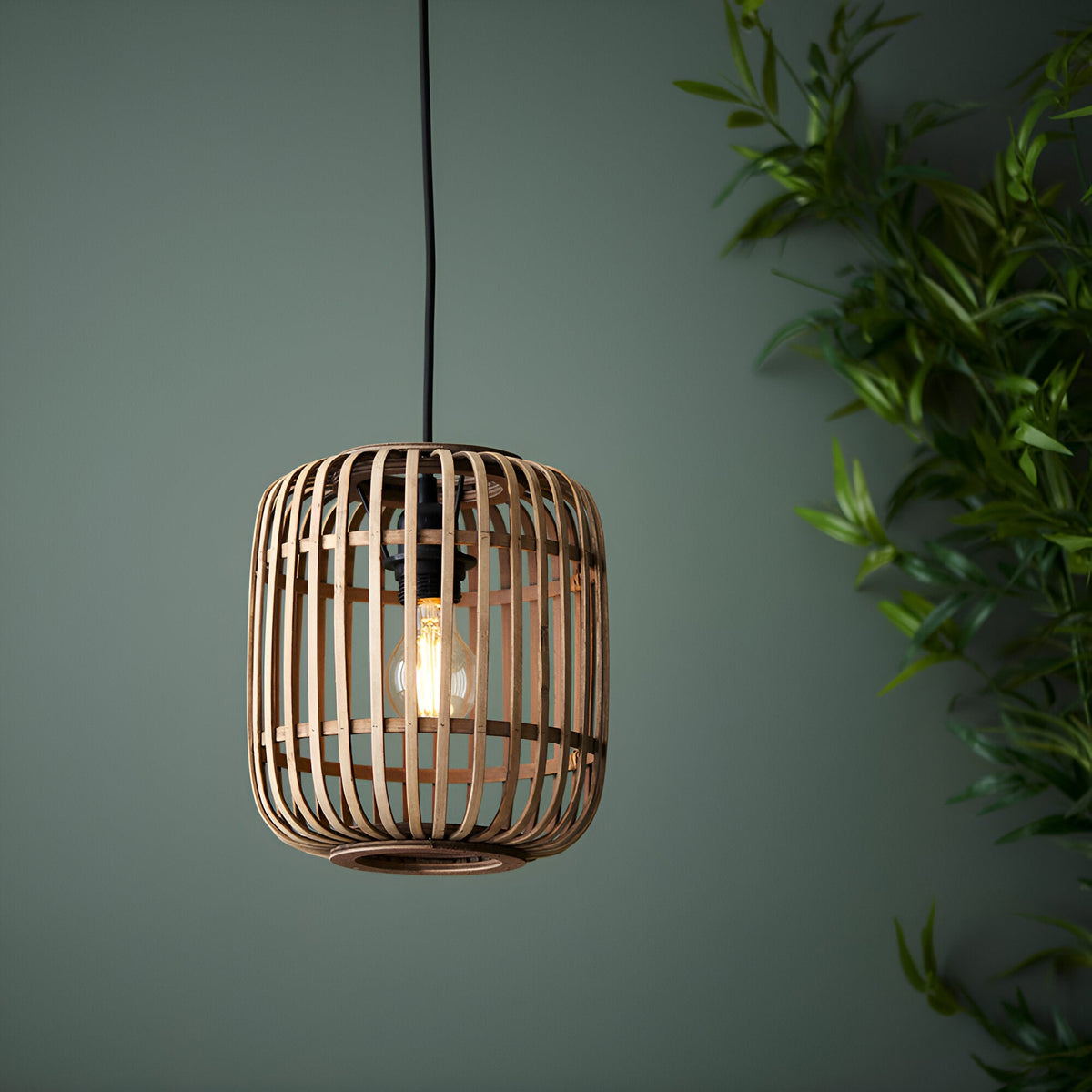 Endon Bracken pendant light with a natural bamboo slatted cage shade and matt black fitting, illuminated by an exposed filament bulb against a green wall.