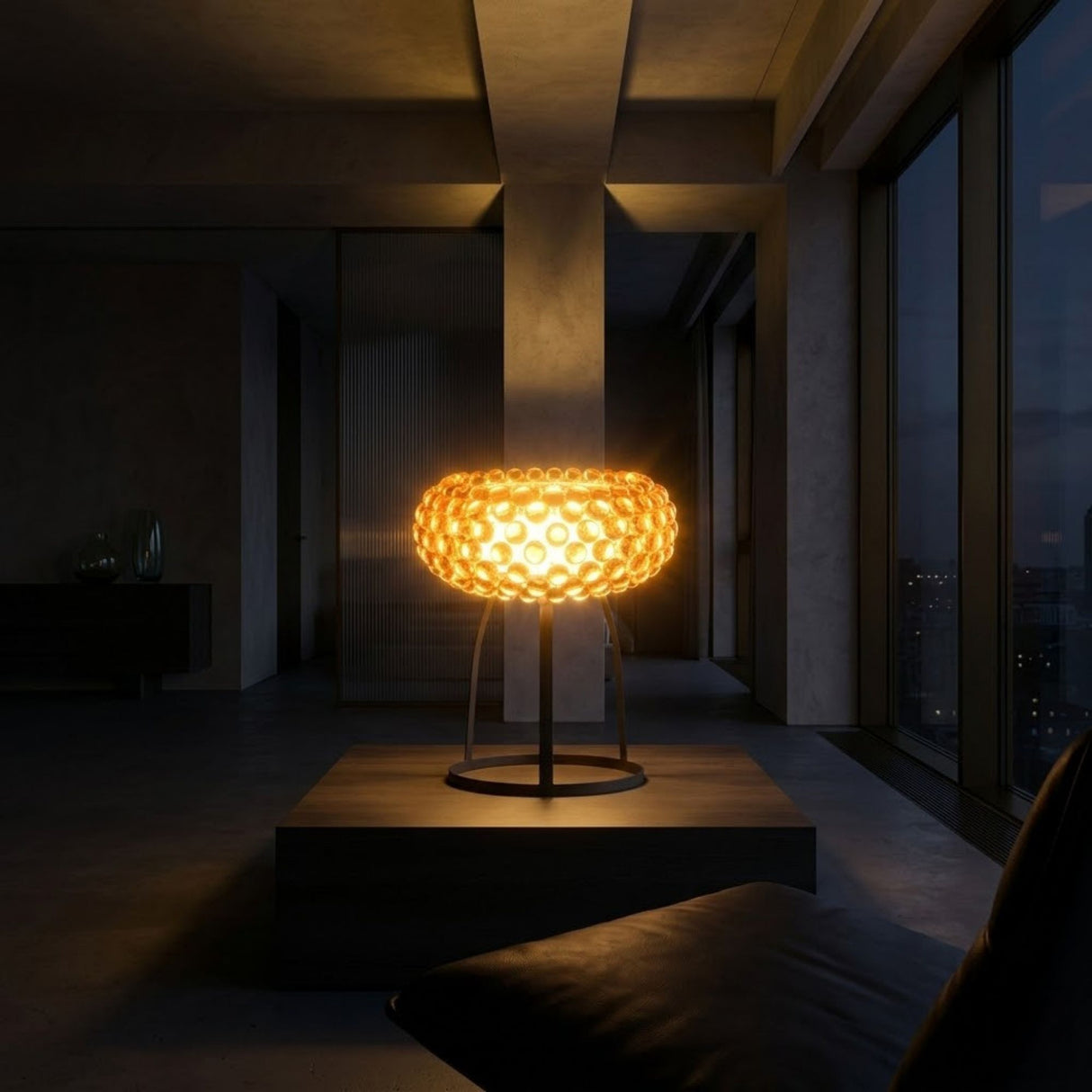 The Briller LED Table Lamp - Gold, featuring a glowing textured circular design, sits on a pedestal in a dimly lit minimalist room with large windows and nighttime city views.