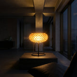 The Briller LED Table Lamp - Gold, featuring a glowing textured circular design, sits on a pedestal in a dimly lit minimalist room with large windows and nighttime city views.