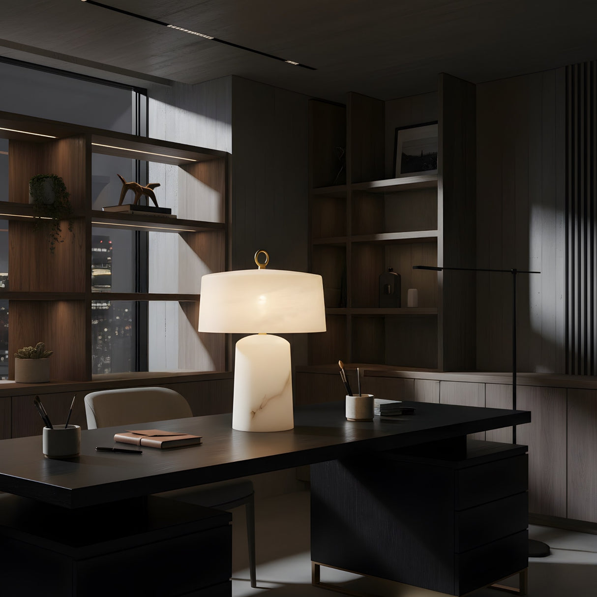A modern, dimly lit office with wooden shelves and a dark desk features the Calyra 3 Light Solid Form Alabaster Table Lamp – Soft White, softly illuminating notebooks and pens as city lights shine through the window in the background.