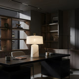 A modern, dimly lit office with wooden shelves and a dark desk features the Calyra 3 Light Solid Form Alabaster Table Lamp – Soft White, softly illuminating notebooks and pens as city lights shine through the window in the background.