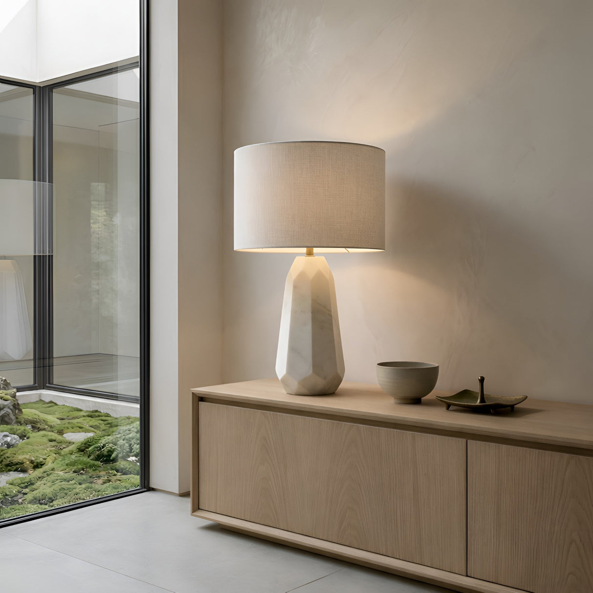 A modern interior features a light wood sideboard with the Averon 1 Light Faceted Alabaster Table Lamp in Brass & Soft White, a ceramic bowl, and a decorative tray. Large windows reveal a lush green garden outside.