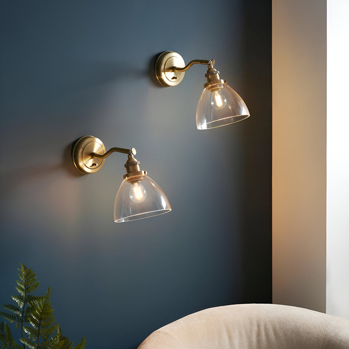 Wentworth Wall Light in antique brass with adjustable knuckle arm and clear glass shade, shown as a pair mounted on a dark blue wall in a living room setting.