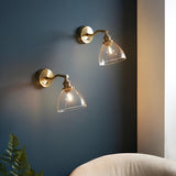 Wentworth Wall Light in antique brass with adjustable knuckle arm and clear glass shade, shown as a pair mounted on a dark blue wall in a living room setting.