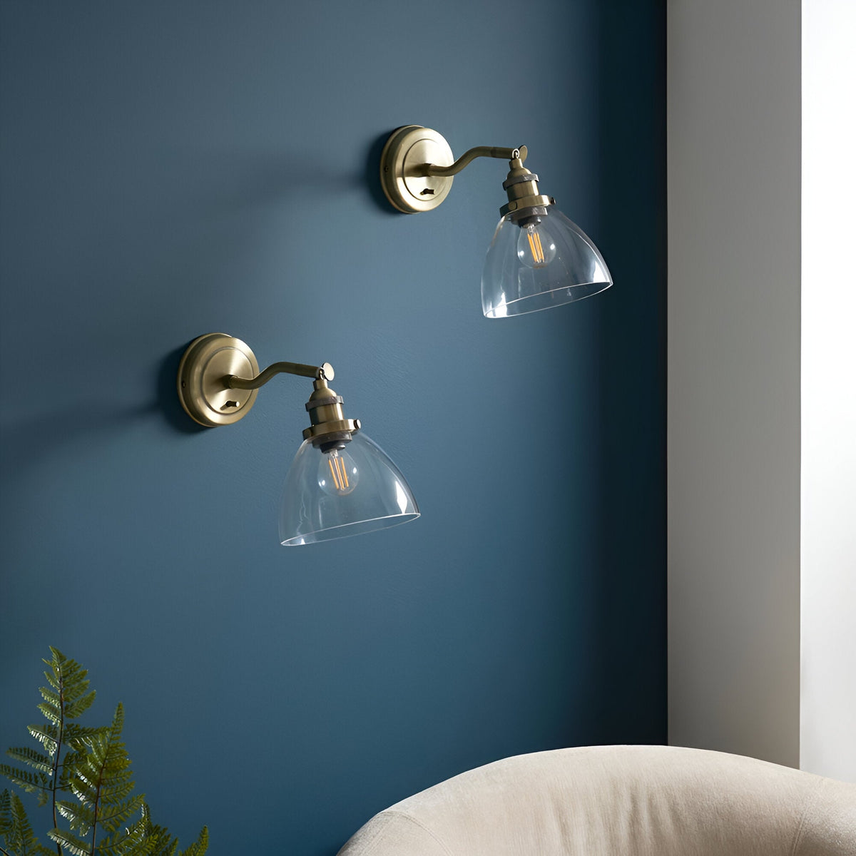 Wentworth Wall Light in antique brass with adjustable knuckle arm and clear glass shade over a filament bulb, shown as a pair on a dark blue wall.