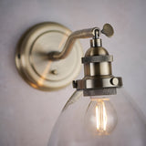 Wentworth Wall Light in antique brass with adjustable knuckle joint, knurled lamp holder and clear glass shade over a filament bulb, mounted on a light wall backplate.