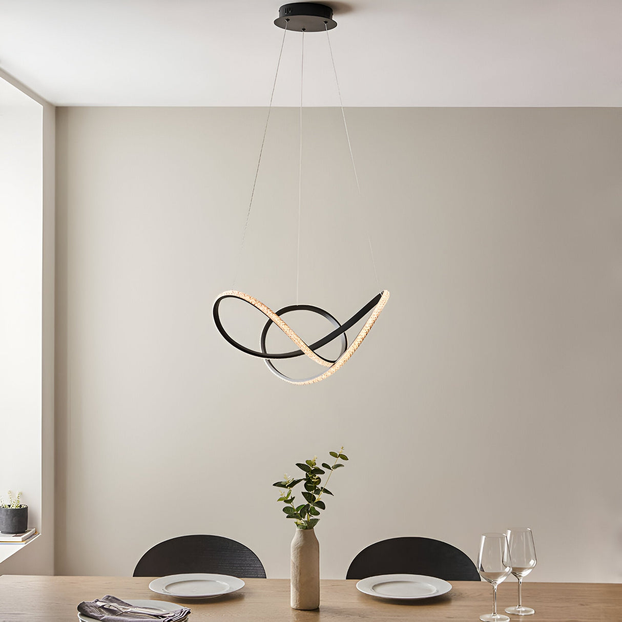 Bramcombe Pendant in matt black with clear faceted acrylic detail, sculptural looped design, hung above a dining table in a neutral-toned room.
