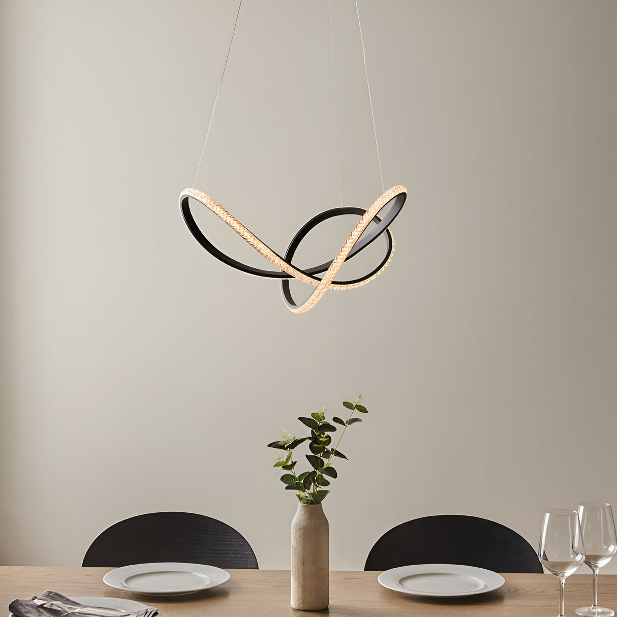 Bramcombe Pendant in matt black with looped, intertwined curves and warm white LED lighting, hung above a dining table in a neutral-toned room.