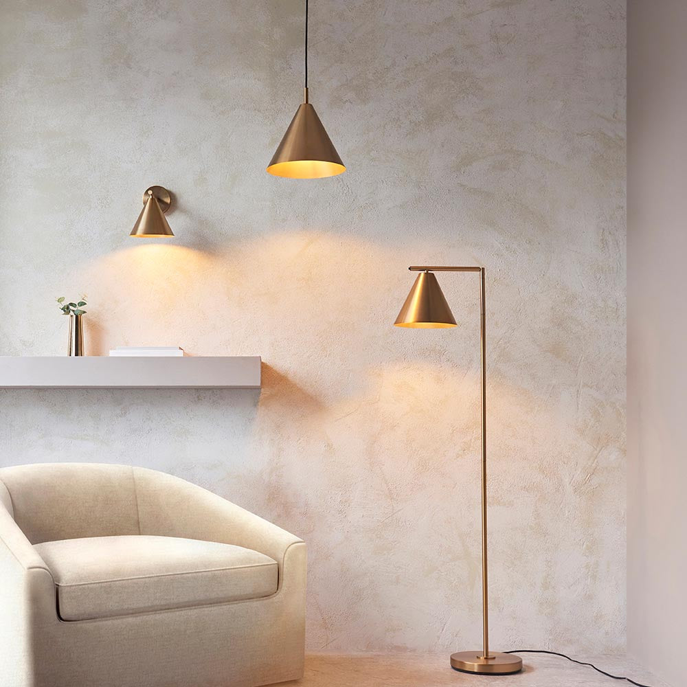 Endon Ashworth brass floor lamp with adjustable conical head beside a cream armchair, shown with matching pendant and wall lights on a textured plaster wall.