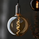 Endon 4W E27 LED filament spiral bulb in amber tinted globe glass on a chrome pendant fitting, glowing warm white against a dark grey backdrop with eucalyptus.