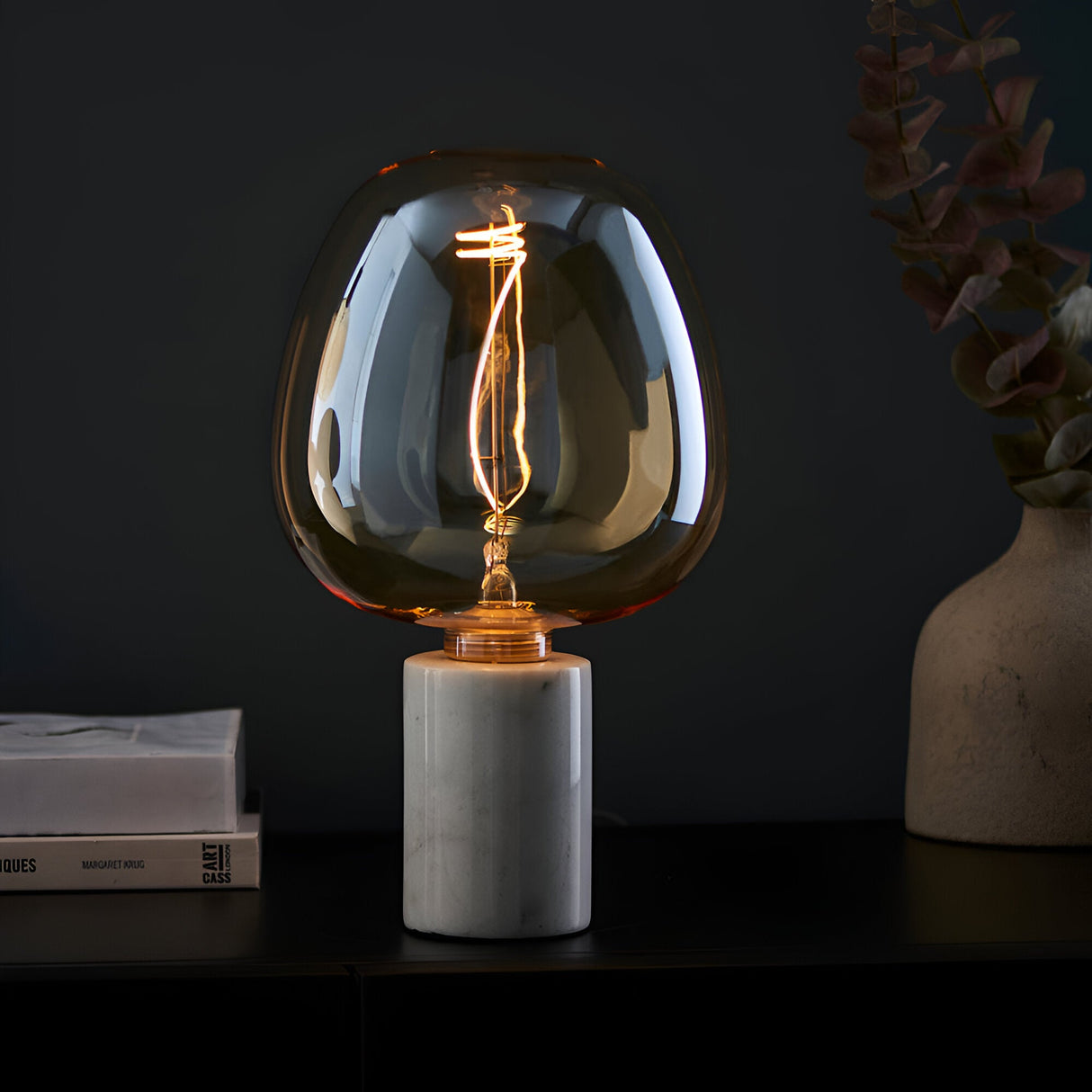 Endon 4W E27 LED filament swirl bulb with amber tinted glass, lit on a marble cylinder holder beside books and a vase in a dark setting.