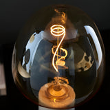 Endon 4W E27 LED filament swirl bulb illuminated in warm amber, showing the curved filament through tinted glass against a dark background.