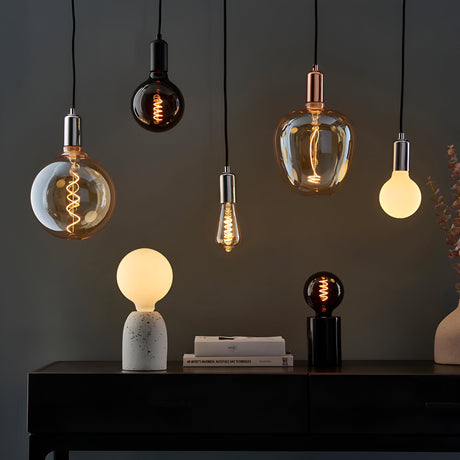 Endon 4W E27 LED filament swirl bulbs in amber and smoked glass hung as pendants above a black console with lit globe table lamps and books.