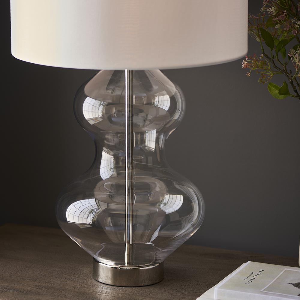 Endon Brockley table lamp with a clear glass hourglass base on a polished nickel foot and white fabric shade, styled on a wood console beside foliage.