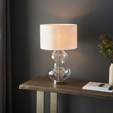 Endon Brockley table lamp with clear glass hourglass base, polished nickel fittings and white drum shade, on a wood console table against a grey wall.