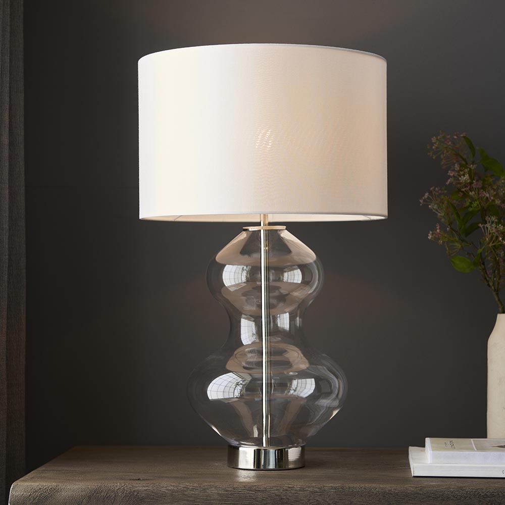 Endon Brockley table lamp with clear glass hourglass base, polished nickel stem and vintage white drum shade, shown lit on a wood console.