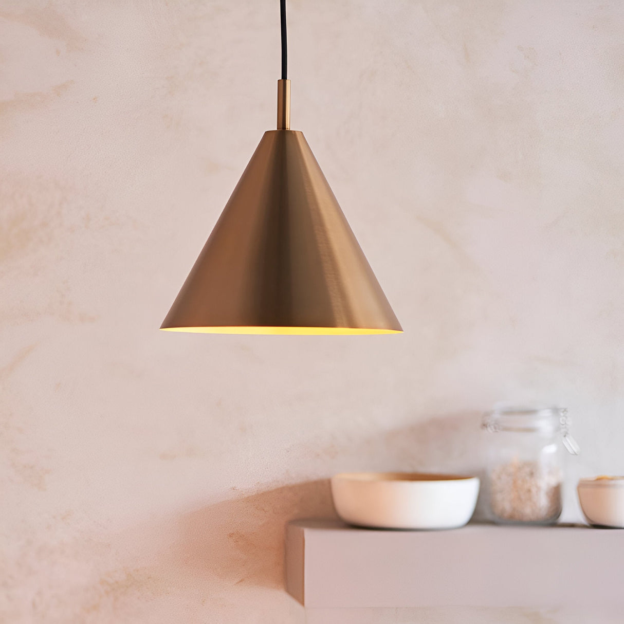 Ashworth Pendant in brass, a cone-shaped single-light fixture with a matching painted inner shade, shown lit above a kitchen shelf with bowls and jars.
