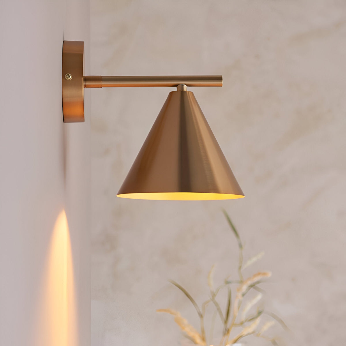 Endon Ashworth brass wall light with adjustable arm and conical shade, mounted on a light wall beside dried grasses, casting a warm glow.