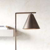 Ashworth Floor Lamp in bronze metal with adjustable arm and cone-shaped shade, shown beside a shelf of books against a light neutral wall.