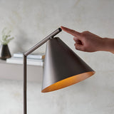 Ashworth Floor Lamp in bronze metal with a conical shade and adjustable angled arm, shown with a hand touching the push switch near a console table.