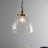 Wentworth Pendant in brass with clear glass dome shade, knurled lampholder, and chain suspension, shown lit against a plain grey wall.