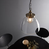 Wentworth Pendant in brass, industrial-style dome shade in clear glass with visible filament bulb, hung on a chain above a dark round table.