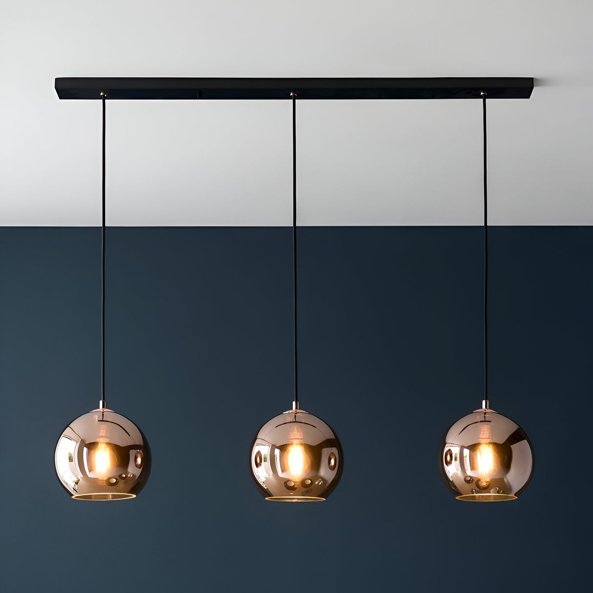 Endon Ashbourne 3 light linear pendant with copper mirrored glass globe shades on a matt black bar, mounted against a two-tone white and navy wall.