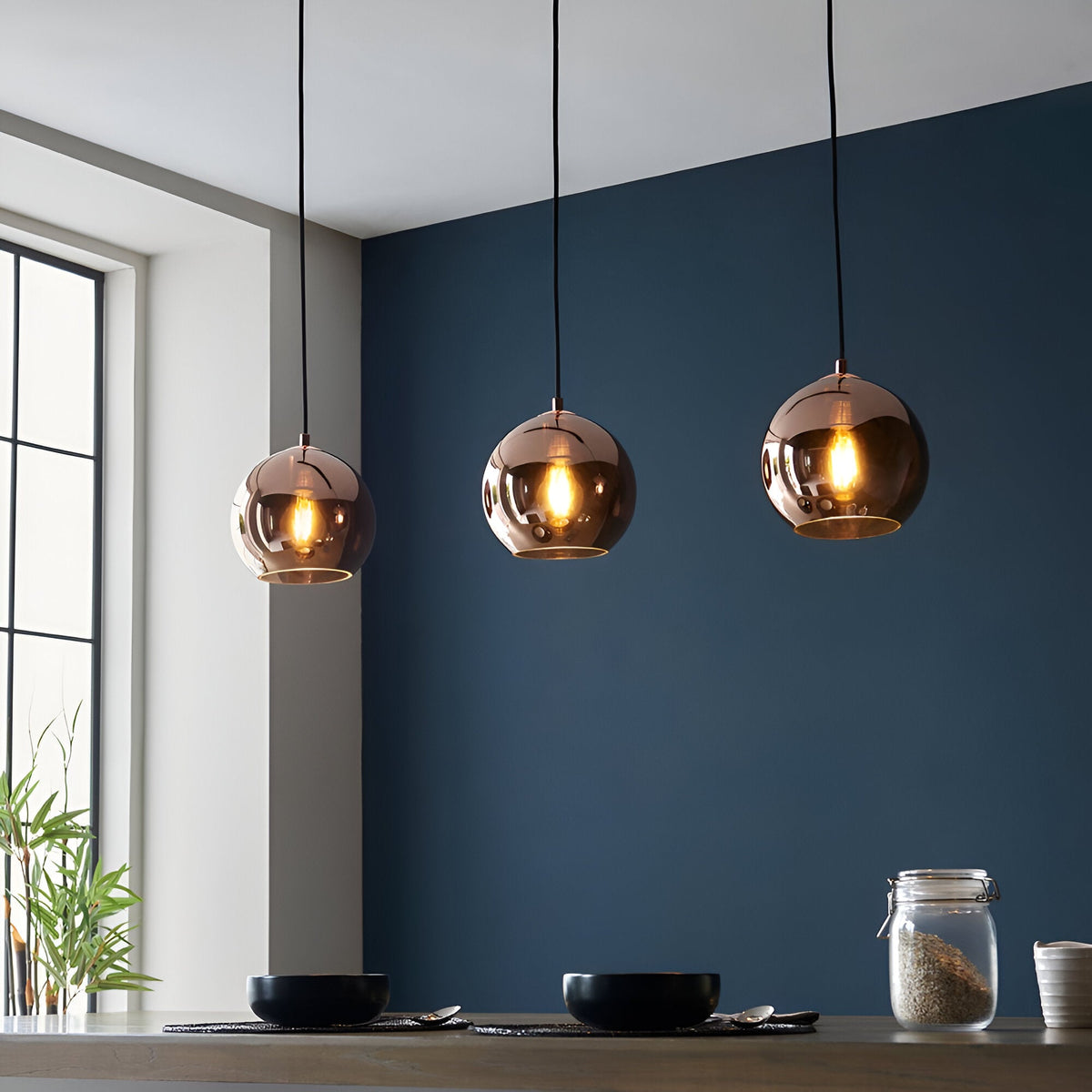 Endon Ashbourne 3 light linear pendant with copper mirrored glass globes on black cables, illuminated above a kitchen counter against a deep blue wall.