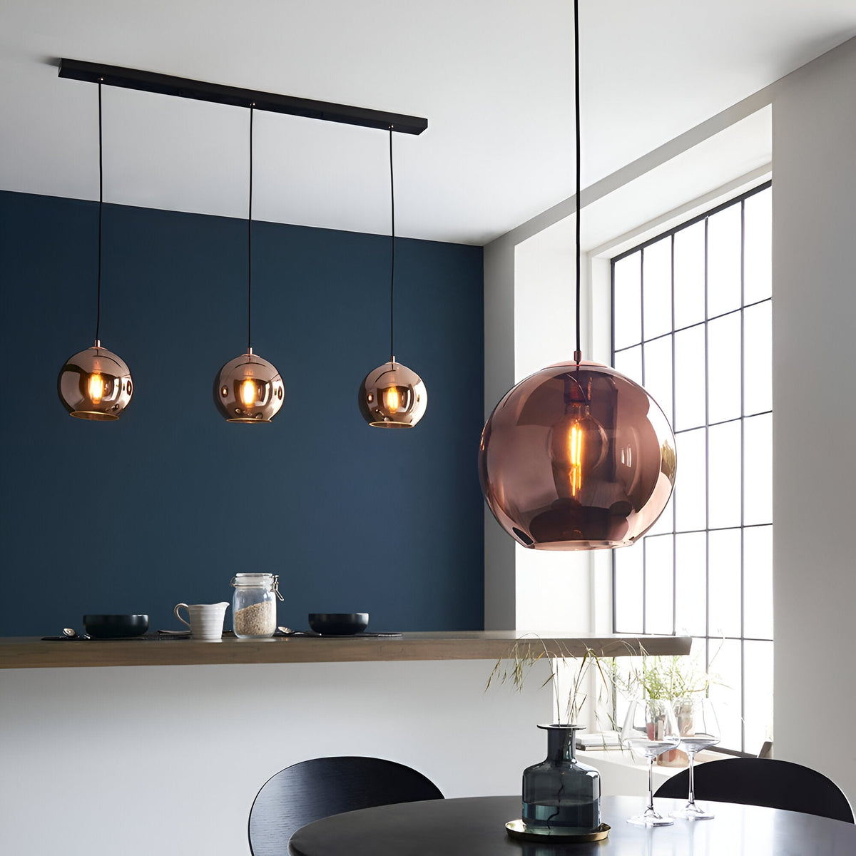 Endon Ashbourne 3 light linear pendant with copper mirrored glass globes on matt black bar, shown beside a larger copper globe pendant in a dining space.