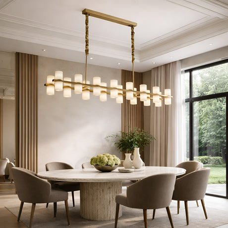 A modern dining room with a round marble table and gray chairs features the Valenro 28 Light Cylindrical Alabaster Linear Suspension Light in soft white & brass, tall beige curtains, and vases with flowers by a window overlooking greenery.