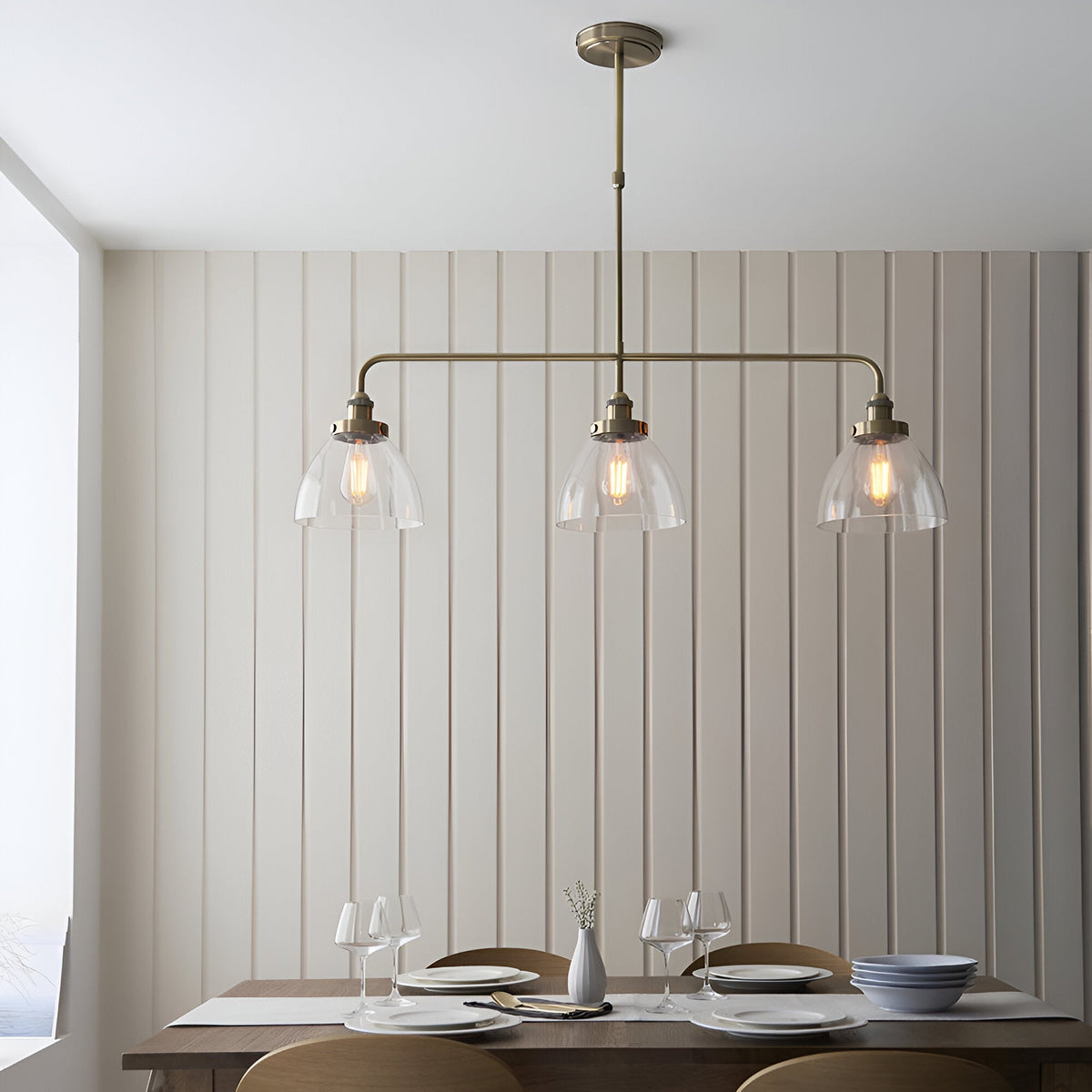 Endon Wentworth 3 light linear stem pendant in antique brass with three clear glass dome shades and exposed filament bulbs, above a dining table.