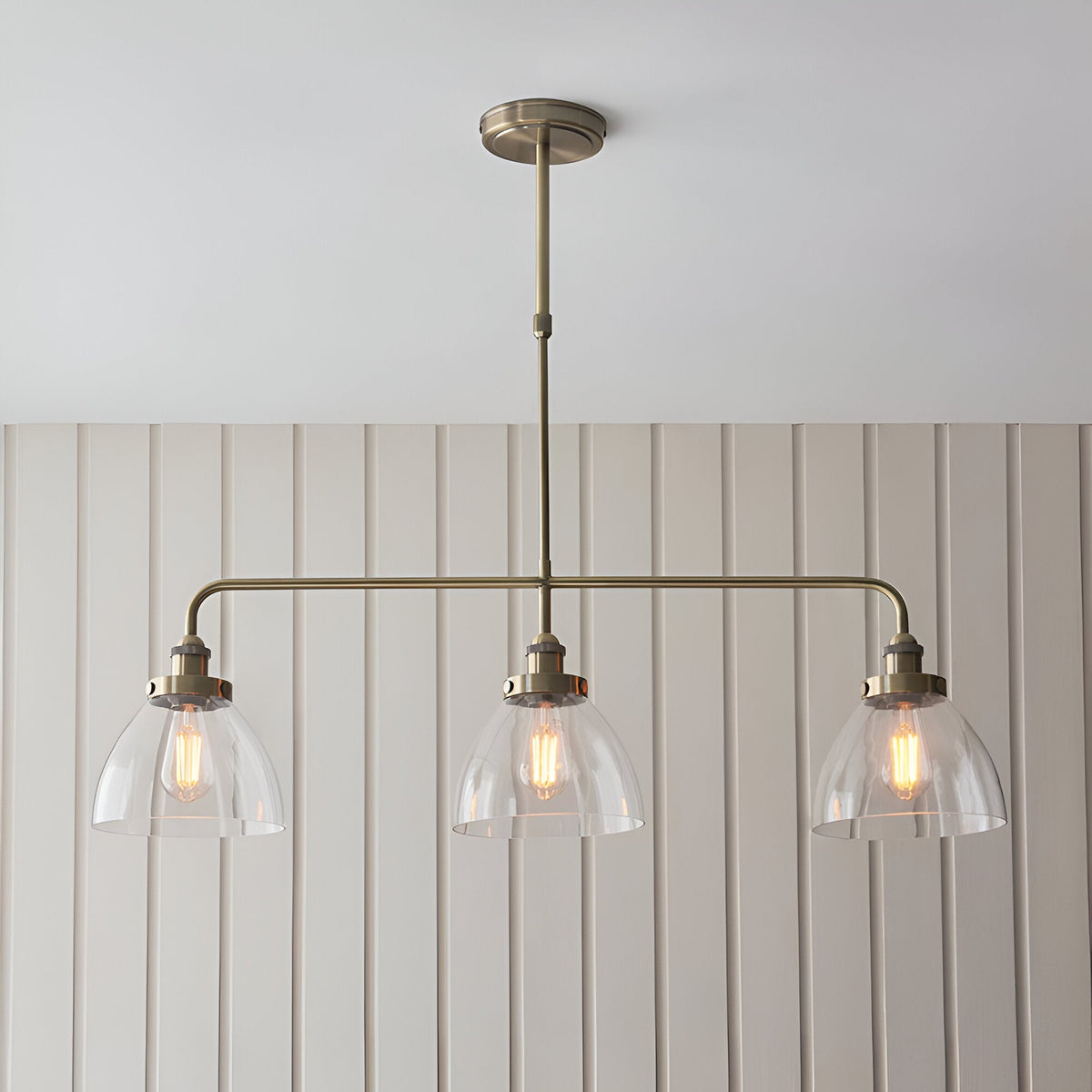 Endon Wentworth 3 light linear stem pendant in antique brass with three clear glass dome shades and exposed filament bulbs, mounted below a panelled wall.