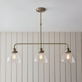 Endon Wentworth 3 light linear stem pendant in antique brass with three clear glass dome shades and exposed filament bulbs, mounted below a panelled wall.