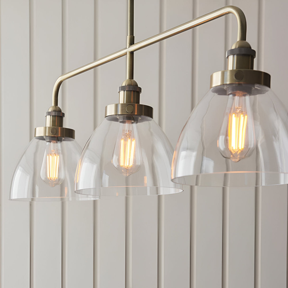Endon Wentworth 3 light linear stem pendant in antique brass with clear glass dome shades and exposed filament E27 lamps, shown against panelled walls.
