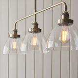Endon Wentworth 3 light linear stem pendant in antique brass with clear glass dome shades and exposed filament E27 lamps, shown against panelled walls.