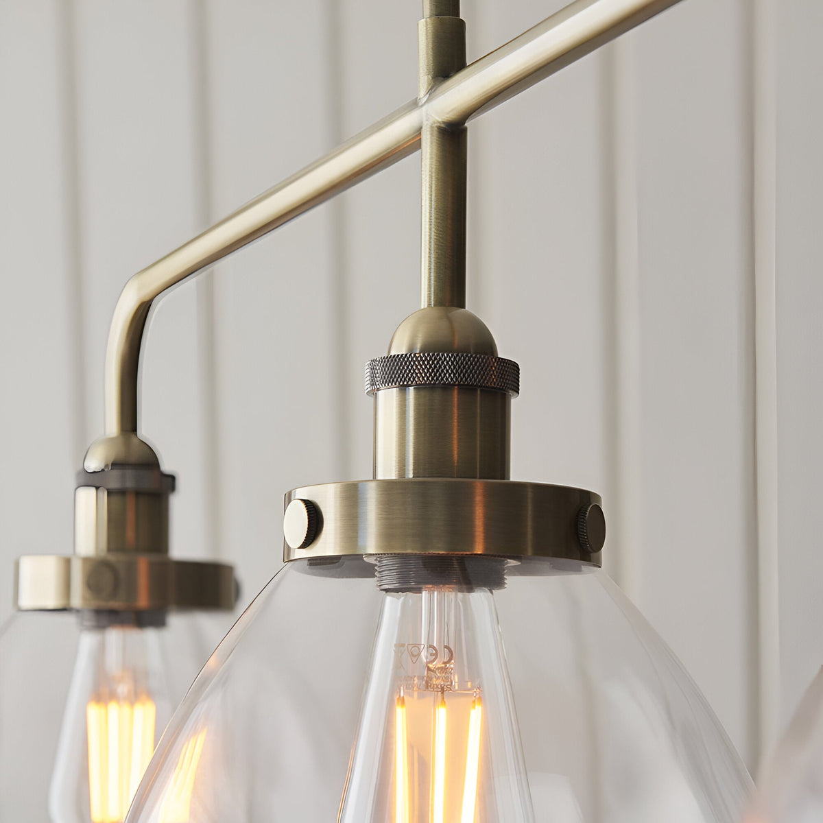 Close-up of Endon Wentworth 3 light linear stem pendant in antique brass, showing knurled lamp holders, clear glass shades and filament bulbs.