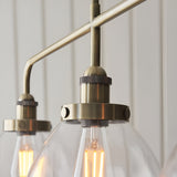 Close-up of Endon Wentworth 3 light linear stem pendant in antique brass, showing knurled lamp holders, clear glass shades and filament bulbs.