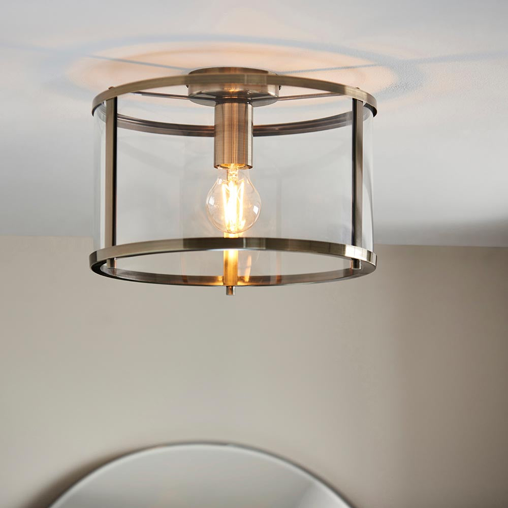 Endon Ellery brass flush ceiling light with cylindrical glass shade and single exposed E27 filament bulb, mounted on a ceiling above a mirror.