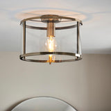 Endon Ellery brass flush ceiling light with cylindrical glass shade and single exposed E27 filament bulb, mounted on a ceiling above a mirror.