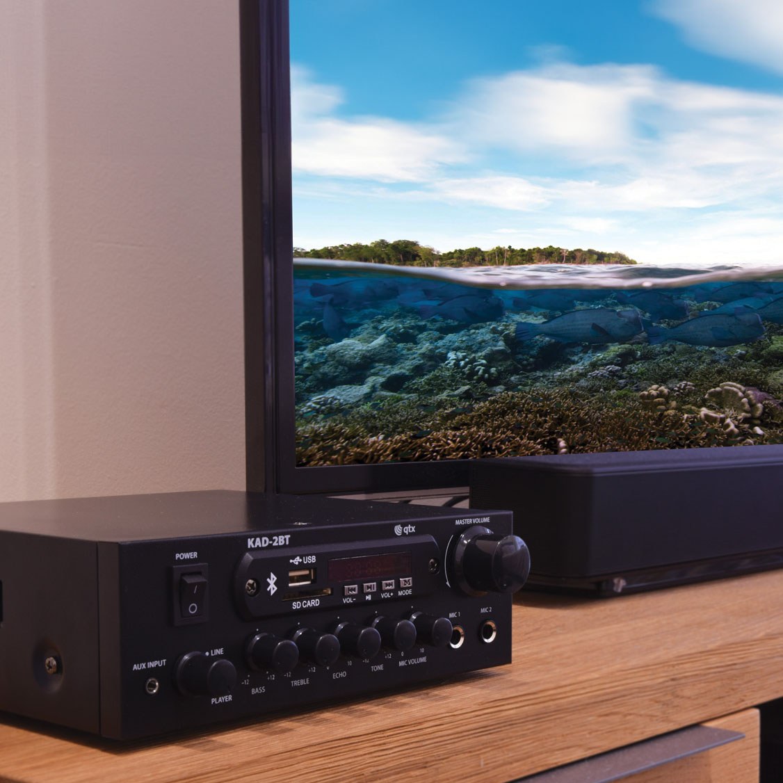 QTX KAD-2BT Bluetooth stereo amplifier in matt black on a wooden shelf, showing USB, SD card slots, master volume knob and mic inputs.