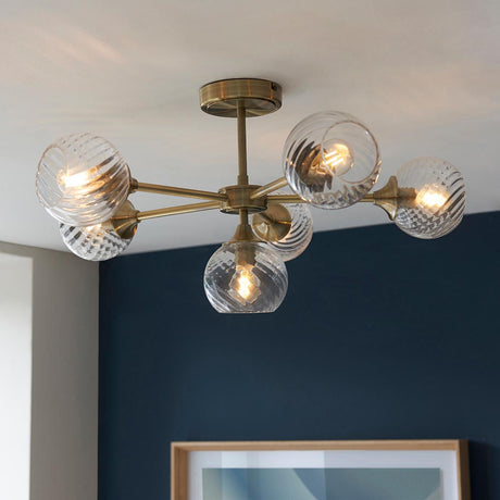 Endon Georgiana 6 light semi-flush ceiling light in brass with ribbed glass globe shades on angled arms, installed against a navy blue wall.