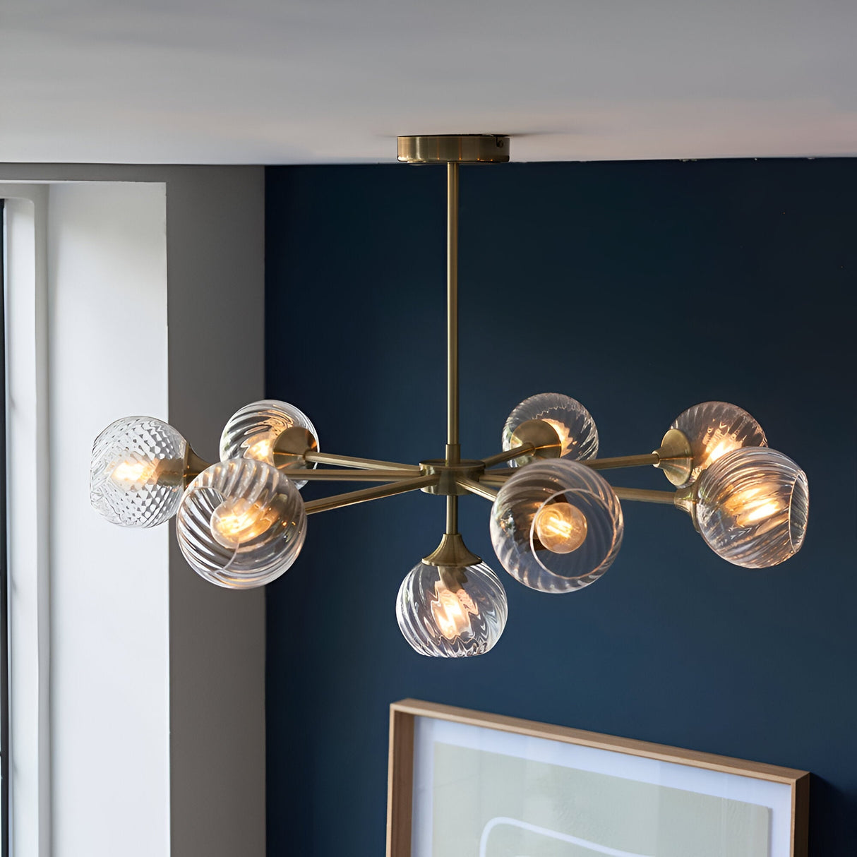 Endon Georgiana 8-light semi-flush pendant in brass with spiral-cut clear glass globe shades, installed against a deep blue wall above a framed print.