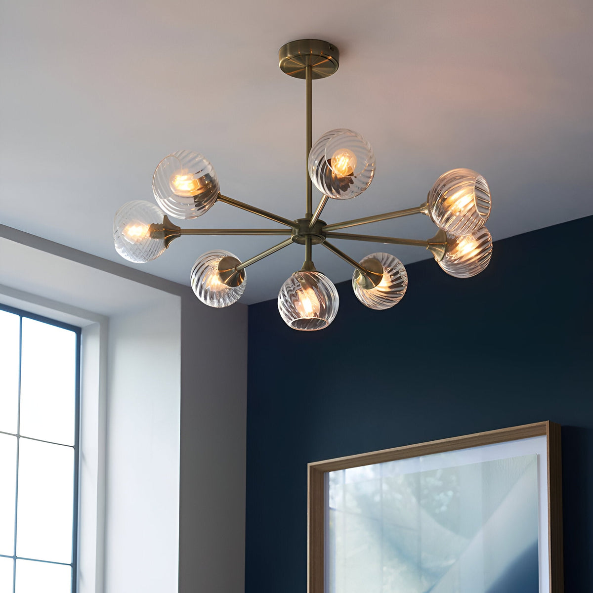 Endon Georgiana 8 light pendant in brass with spiral-cut clear glass globe shades on splayed arms, installed on a white ceiling beside a navy wall.
