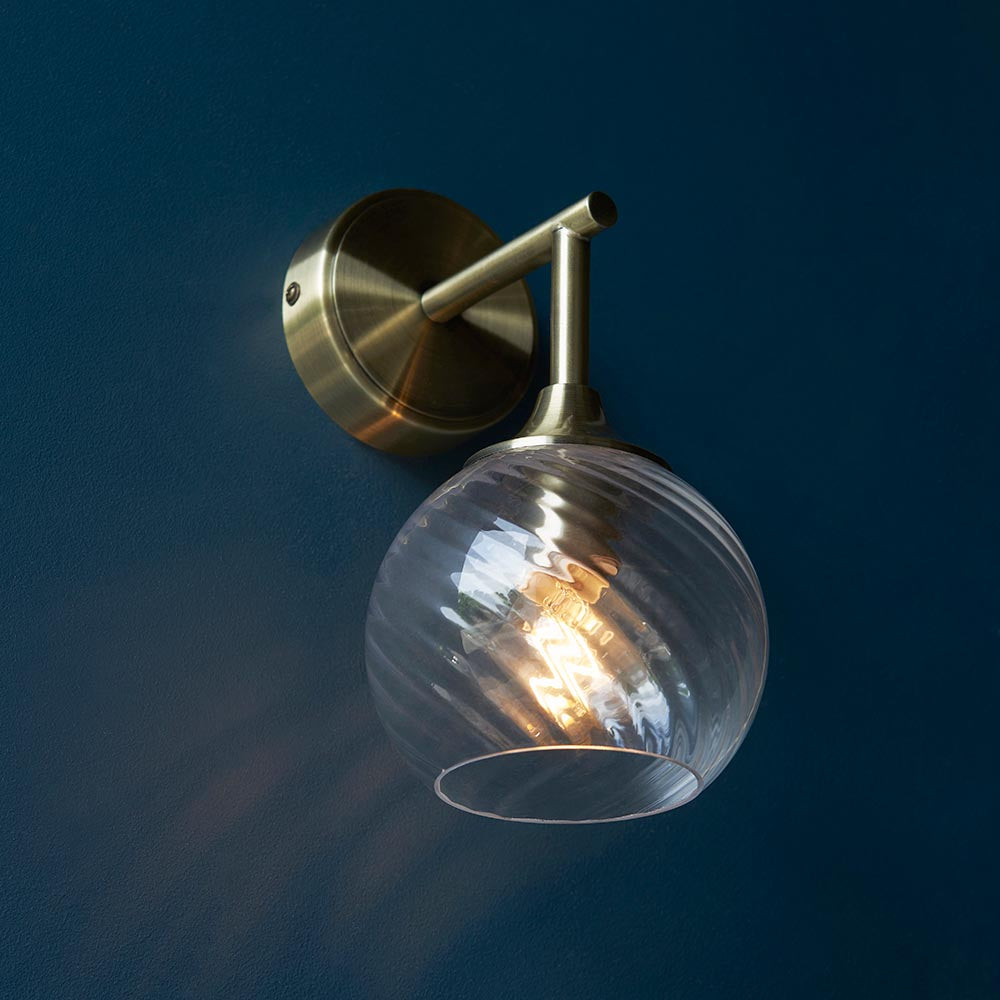 Georgiana Wall Light in brass, with a slim arm holding a spiral-patterned clear glass globe shade, shown lit against a dark blue wall.