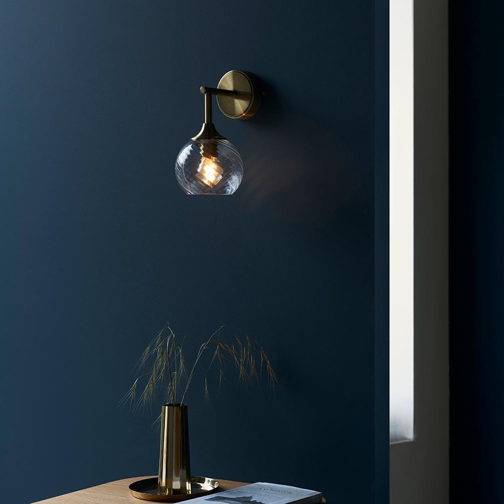 Georgiana Wall Light in brass with a spiral-patterned glass globe shade, mounted on a dark blue wall above a table with a brass vase.