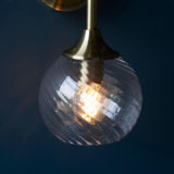 Georgiana Wall Light in brass, single arm with twisted spiral clear glass globe shade, lit bulb visible, set against a dark blue background.